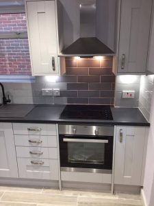 A new kitchen installation featuring grey tiled walls, new integrated cooker with extractor and hob and white kitchen cabinets and cupboards.