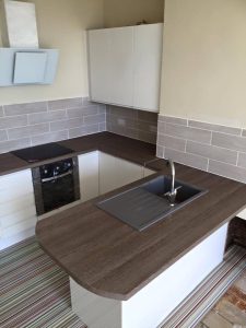 A new kitchen install featuring wooden kitchen worktops, white cabinets, striped vinyl flooring and grey wall tiling.