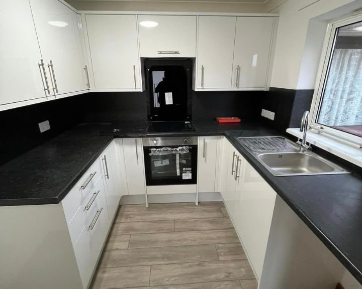 A kitchen design in Basingstoke featuring white kitchen cabinets, wooden vinyl flooring, black countertops and a metal sink.