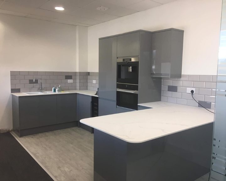 A new commercial kitchen installation featuring grey kitchen cupboards and cabinets, white countertops, wooden vinyl flooring and grey wall tiling.