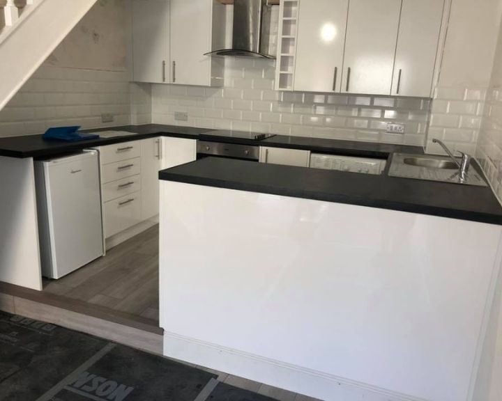 A kitchen under some stairs that has been refurbished with new white kitchen cabinets, vinyl wooden flooring and white kitchen wall tiles.